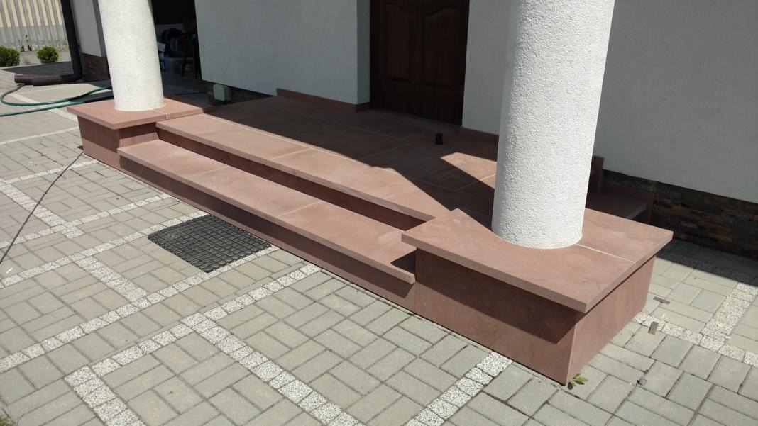 Trzy stopnie wejściowe z piaskowca prowadzące do drzwi budynku, z kolumną po prawej stronie, ułożone na kostce brukowej.