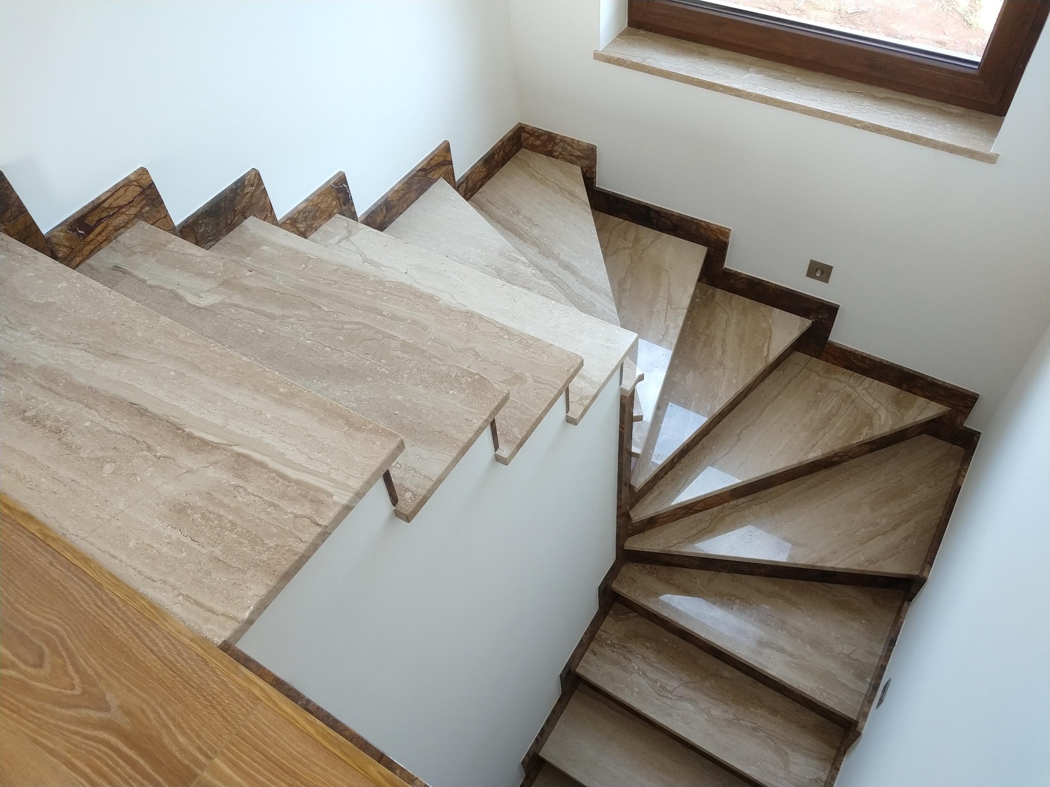 Spiralne schody wewnętrzne z beżowego trawertynu z ciemniejszymi krawędziami, widok z góry, oświetlenie dzienne, widoczny fragment drewnianej podłogi i parapet okienny.