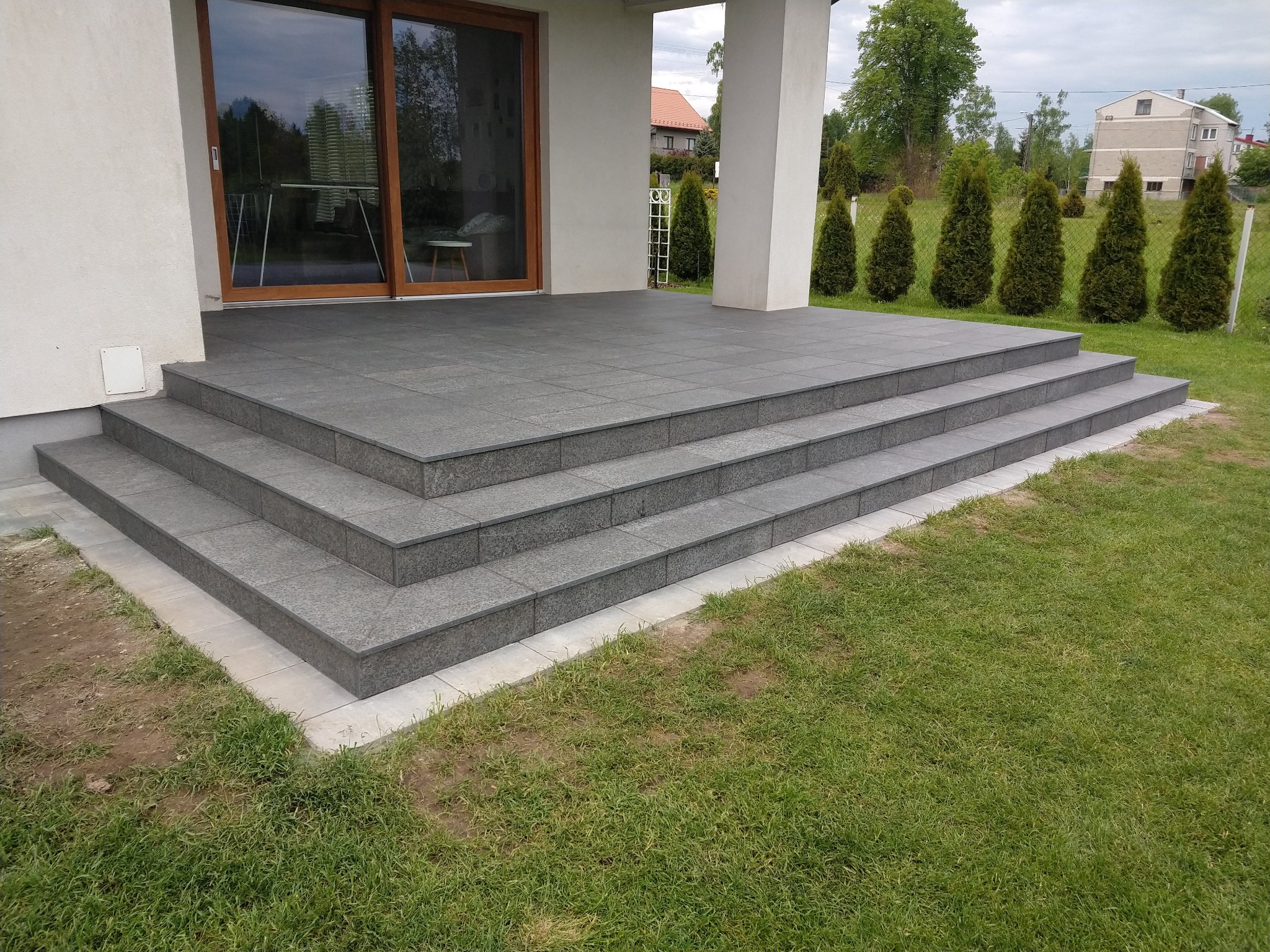 Zewnętrzne schody kamienne prowadzące na taras, wyłożone szarymi płytami, z widokiem na trawnik i tujowe szpalery.