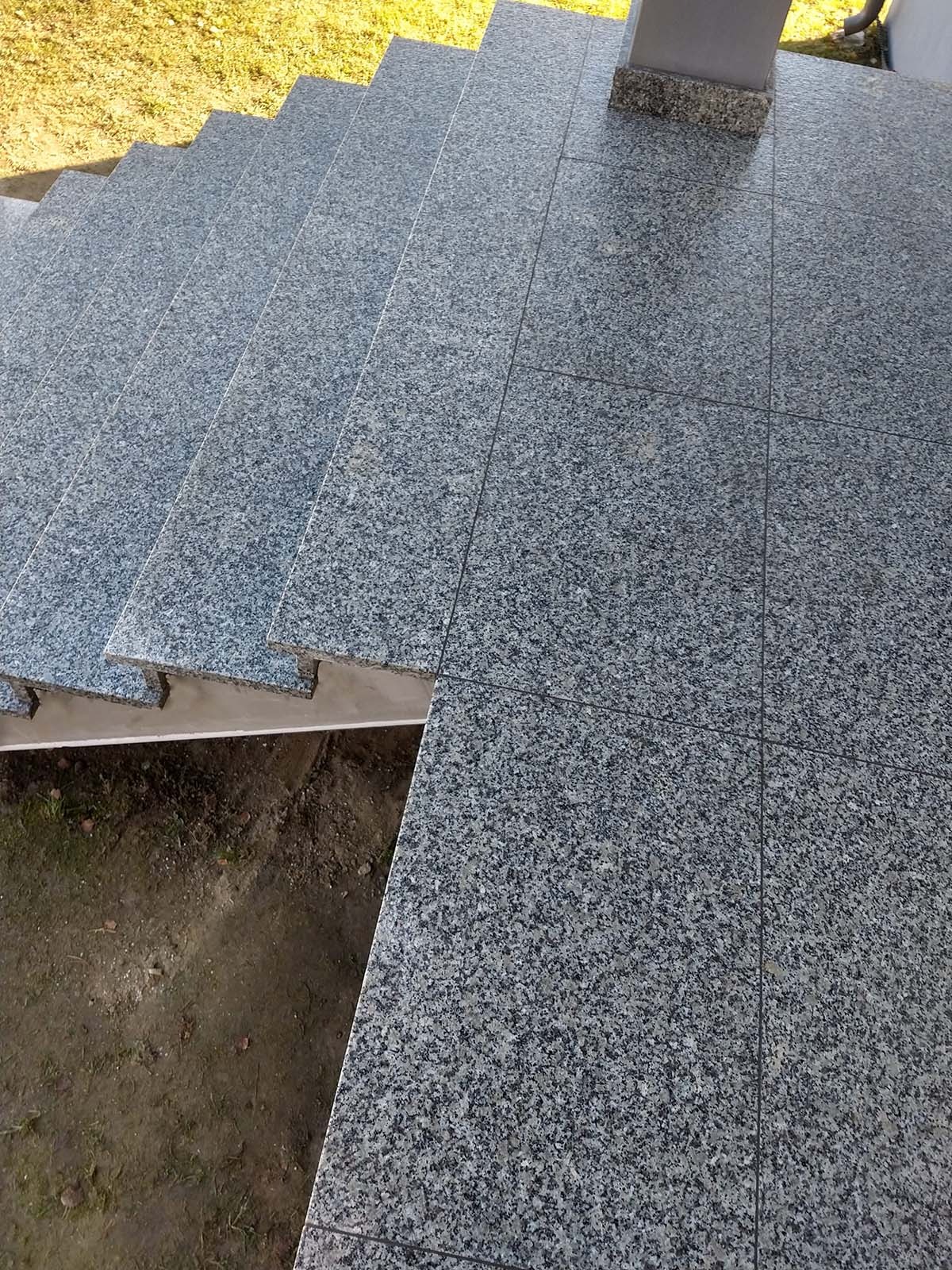 Zewnętrzne schody wyłożone szarymi, kwadratowymi płytami z drobnoziarnistego kamienia naturalnego, widoczne niedokończone podłoże pod schodami.