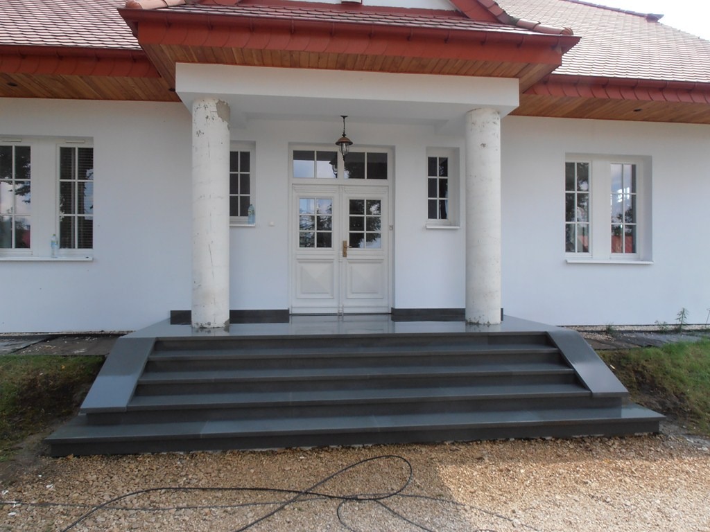 Wejście do domu z ciemnoszarymi, prostymi stopniami i białymi kolumnami, zakończone podestem przed drzwiami.