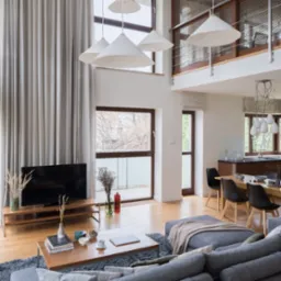 Jasnoszara, długa zasłona w przestronnym salonie z wysokim sufitem i dużymi oknami, widoczne także rolety rzymskie na oknach w kuchni.