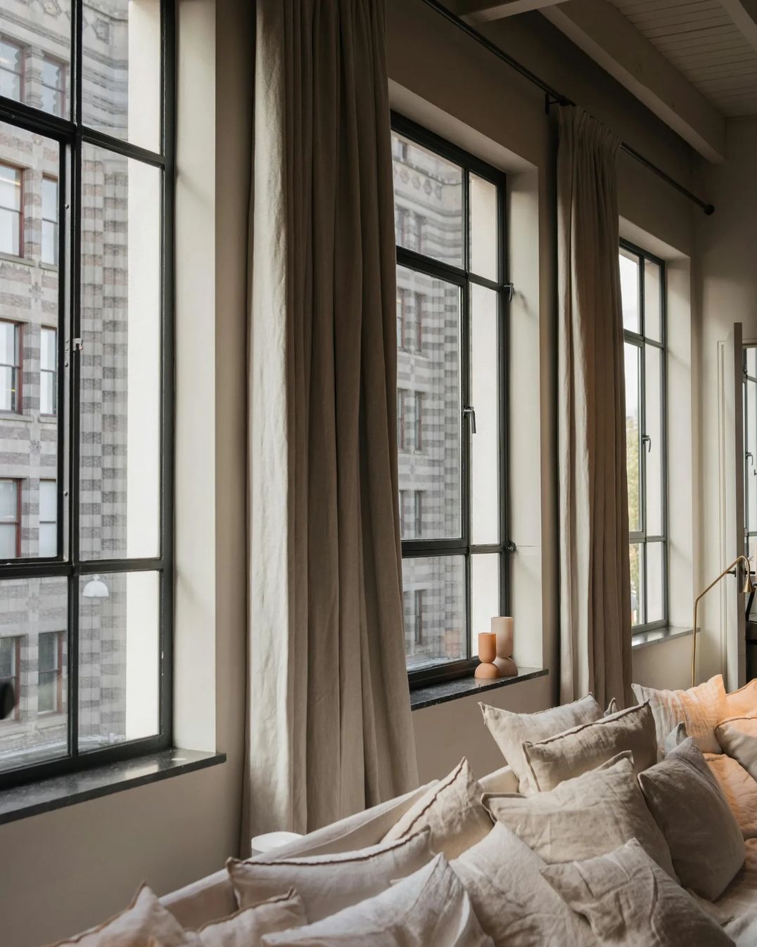 Jasne, lniane zasłony na czarnych karniszach w loftowym wnętrzu z widokiem na budynek z cegły, miękkie poduszki w odcieniach beżu na pierwszym planie.
