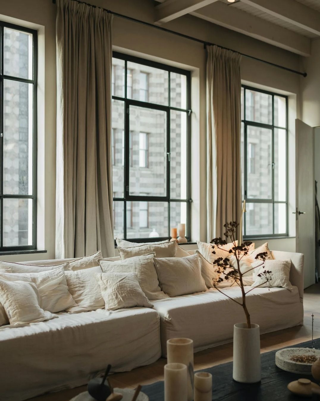 Jasne, lniane zasłony w minimalistycznym salonie z wysokimi oknami i kremową sofą z licznymi poduszkami w odcieniach bieli i beżu, dekoracyjna gałązka w wazonie na stoliku kawowym.