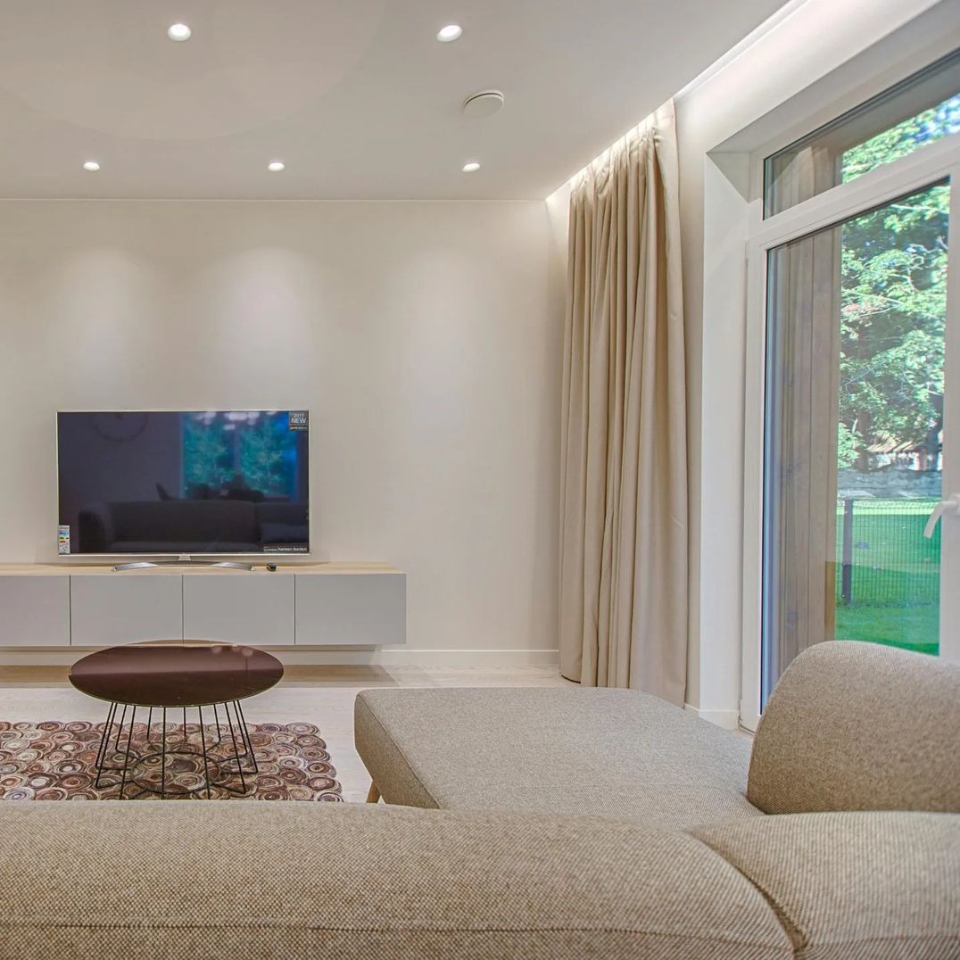 Jasne, minimalistyczne wnętrze z beżowymi zasłonami, telewizorem na niskiej szafce i częściowo widoczną kanapą w podobnym odcieniu.