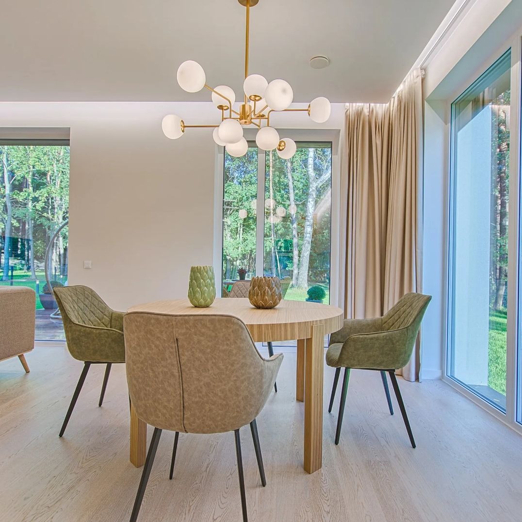 Jasne wnętrze jadalni z okrągłym stołem, czterema krzesłami w odcieniach zieleni i beżu, nowoczesnym żyrandolem z kulistymi kloszami oraz beżowymi zasłonami przy oknach z widokiem na zieleń.