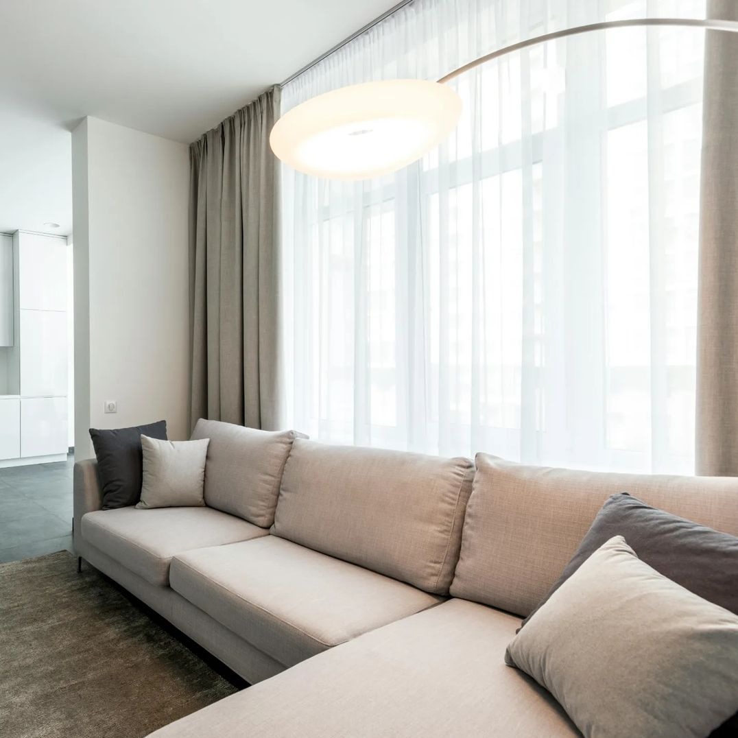 Jasny salon z beżową sofą, szarymi poduszkami i długimi, szarymi zasłonami, przez które prześwituje biała firana, oświetlony lampą łukową