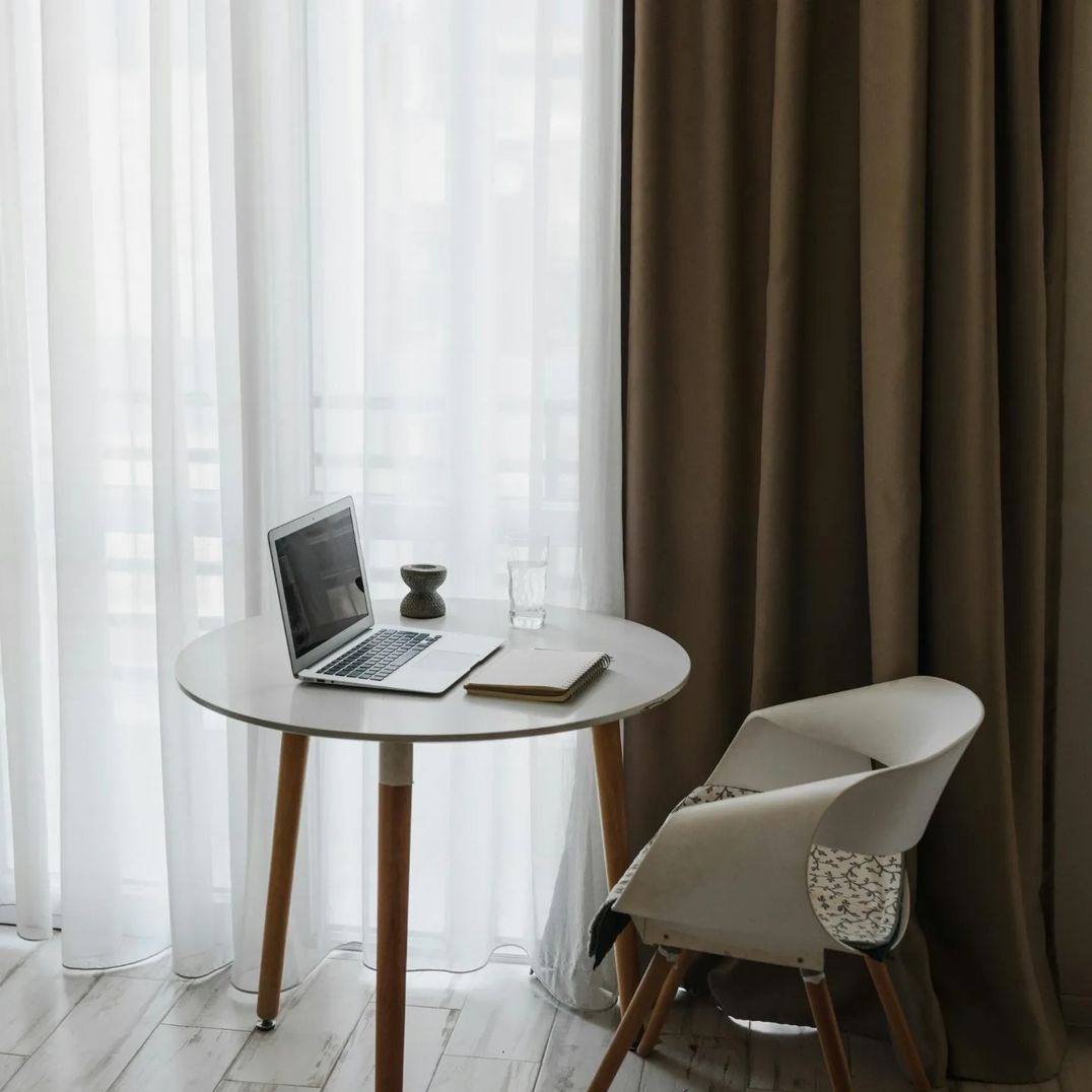 Minimalistyczne wnętrze z okrągłym białym stolikiem, laptopem, szklanką wody i notatnikiem na tle białych, delikatnych firan i brązowych zasłon.
