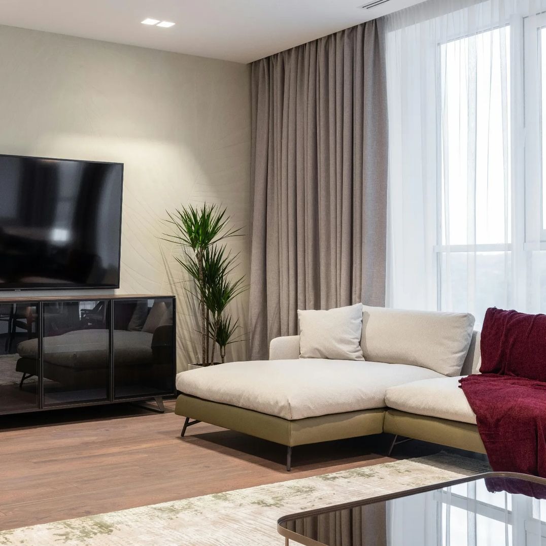 Beżowe, długie zasłony w salonie z narożną kanapą, telewizorem i rośliną doniczkową.
