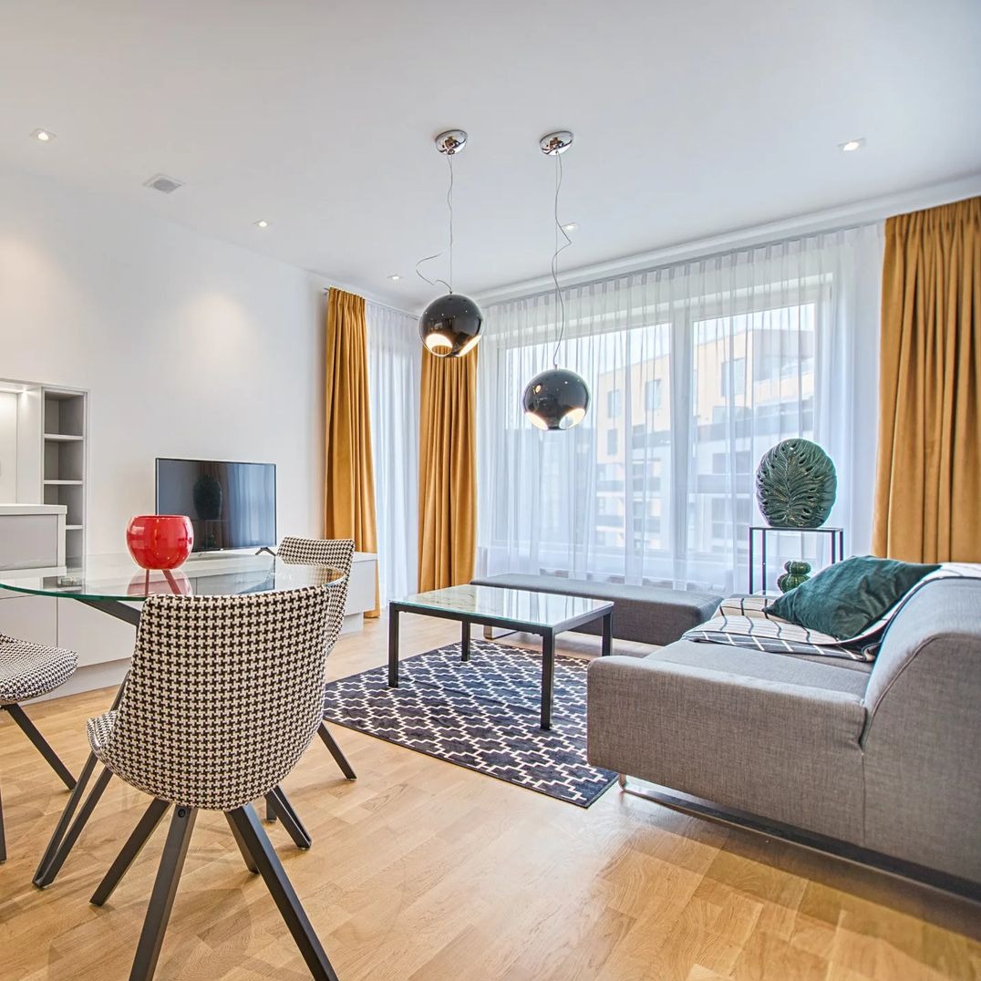 Jasne wnętrze salonu z żółtymi zasłonami, przezroczystymi firanami, szarą sofą, szklanym stołem i krzesłami w pepitkę, oświetlone wiszącymi lampami.