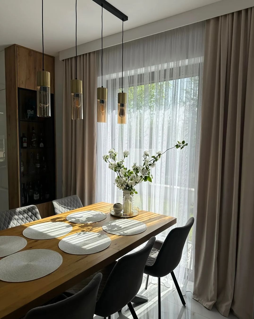 Beżowe, zaciemniające zasłony w nowoczesnej jadalni z drewnianym stołem i bukietem białych kwiatów, wiszące lampy z bursztynowymi kloszami.