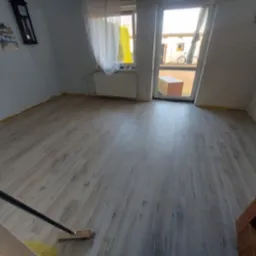 Pokój po położeniu jasnych paneli podłogowych z widoczną fakturą drewna, listwy przypodłogowe jeszcze nie zamontowane, widoczna szczotka do zamiatania.