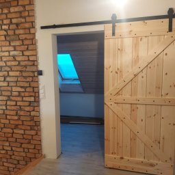 RAFI - BUD RAFAŁ ROMANOWSKI - Drewniane drzwi przesuwne w stylu rustykalnym, zamontowane na czarnej szynie, z widoczną ścianą z czerwonej cegły i oknem dachowym w tle.