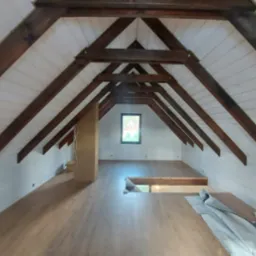 Poddasze po remoncie z drewnianą konstrukcją dachu, wykończone drewnianą boazerią i panelami podłogowymi, widoczne okno dachowe i pionowa zabudowa z przewodami elektrycznymi.