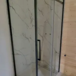 Kabina prysznicowa z przesuwnymi drzwiami w czarnej ramie, zamontowana w łazience z wykończeniem ścian imitującym marmur i boazerią z jasnego drewna.