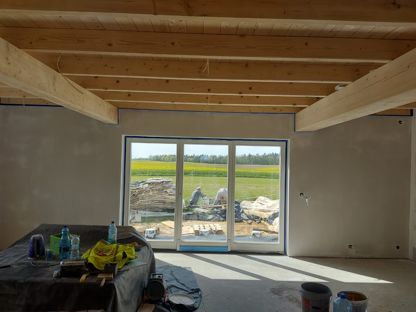 Wnętrze w trakcie prac wykończeniowych: surowy strop z drewnianych belek, ściany przygotowane do malowania z widocznymi puszkami elektrycznymi, duże okno tarasowe z widokiem na pole rzepaku...