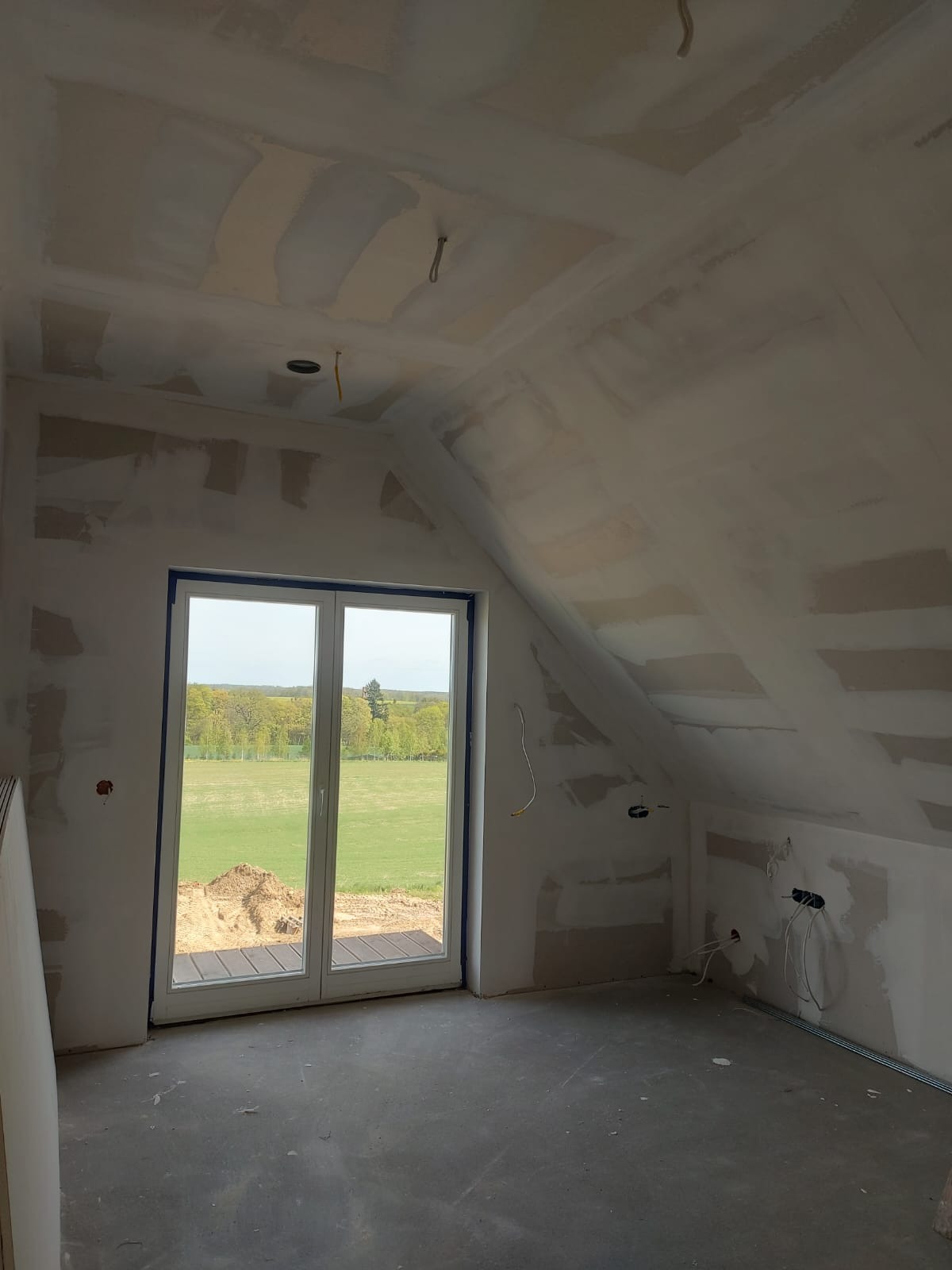 Poddasze w trakcie prac wykończeniowych, widoczne ślady szpachlowania na ścianach i suficie, instalacja elektryczna wyprowadzona ze ściany, okno balkonowe z widokiem na zieleń.