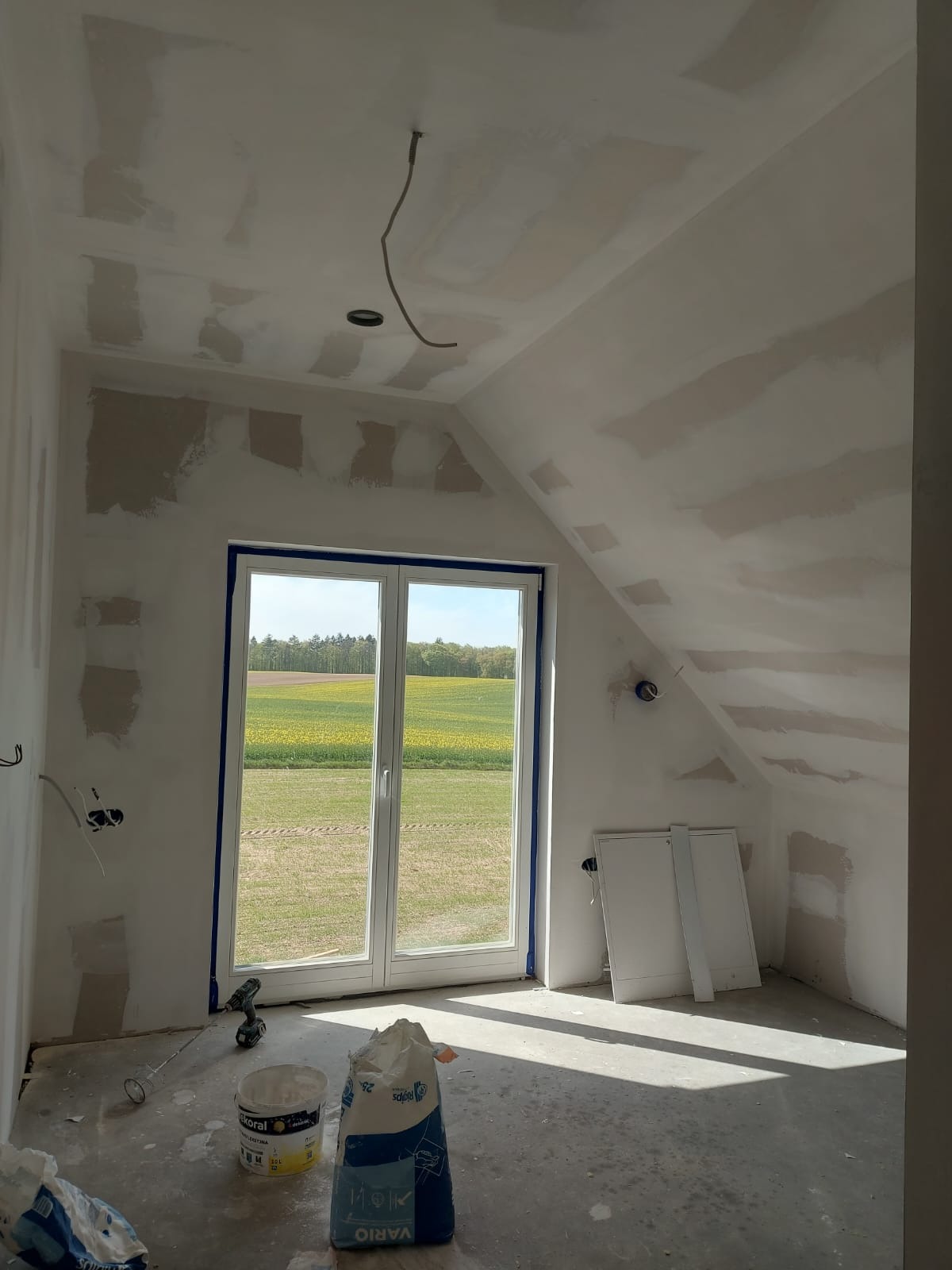 Poddasze w trakcie prac wykończeniowych: ściany i sufit po szpachlowaniu, widoczne worki z zaprawą, okno zabezpieczone taśmą malarską, widok na pola za oknem.