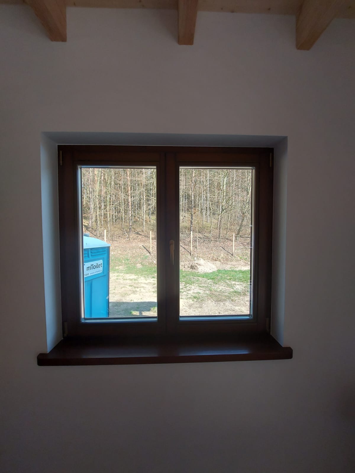 Nowe, brązowe okno PCV z widokiem na zewnątrz, gdzie widać las i przenośną toaletę. Sufit wykończony drewnianymi belkami, ściany gładkie, pomalowane na biało.