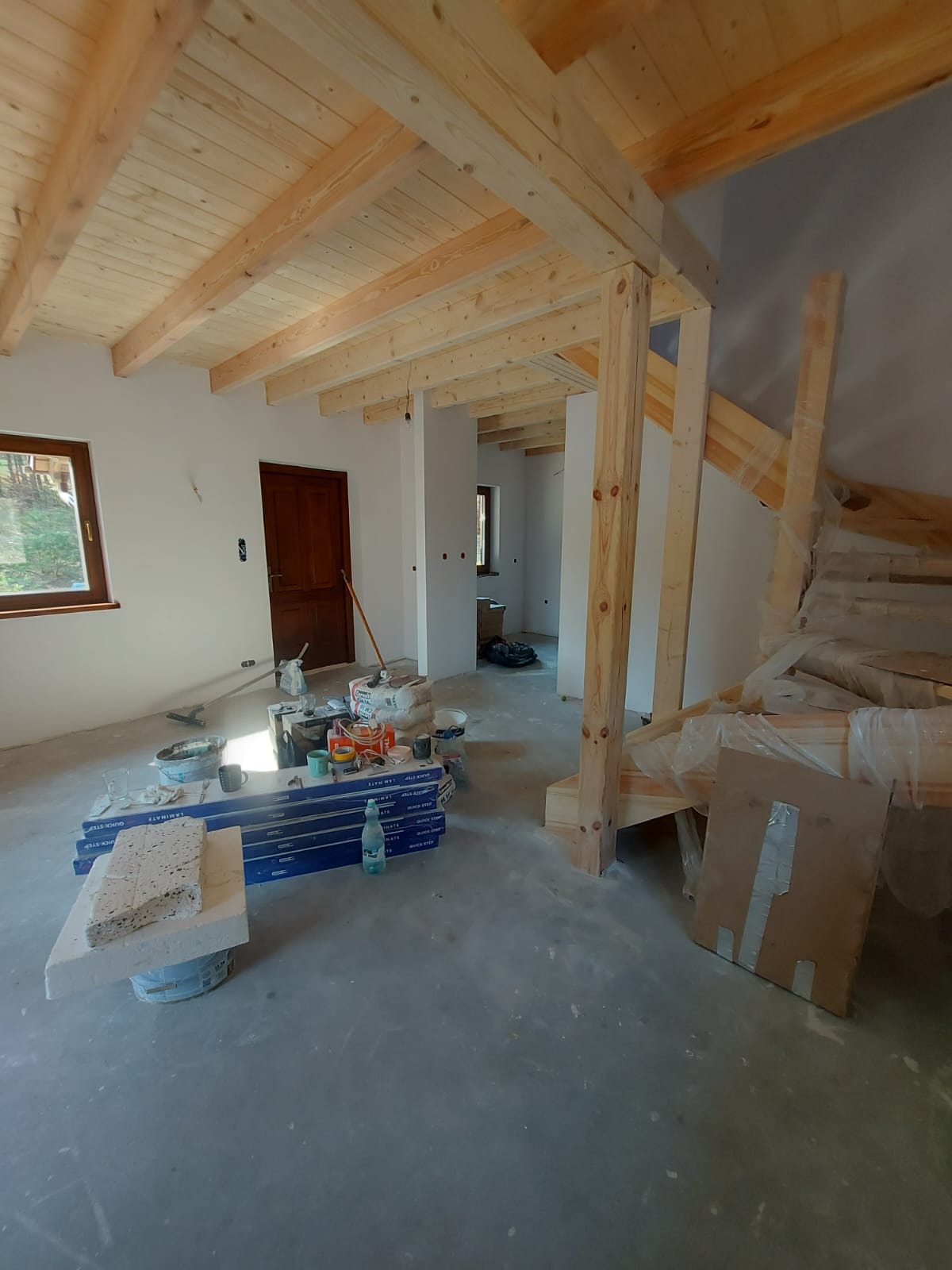 Wnętrze poddasza w trakcie prac wykończeniowych; widoczne drewniane belki stropowe, surowe ściany, podłoga wylewka, okno z widokiem na zewnątrz, materiały budowlane i narzędzia do układania podłogi.