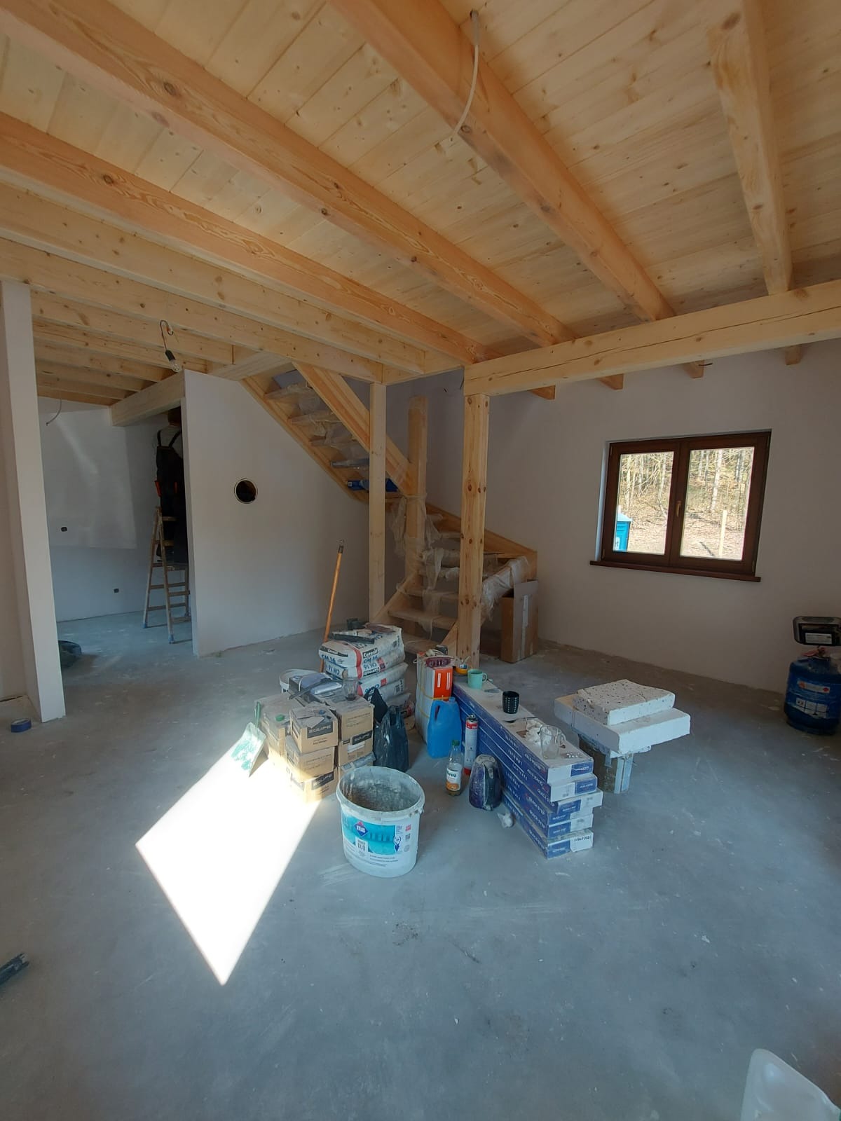 Wnętrze w trakcie prac wykończeniowych: surowy sufit z drewnianych belek, nieotynkowane ściany, drewniane schody w folii ochronnej, materiały budowlane na podłodze, w tle drabina i sylwetka osoby.