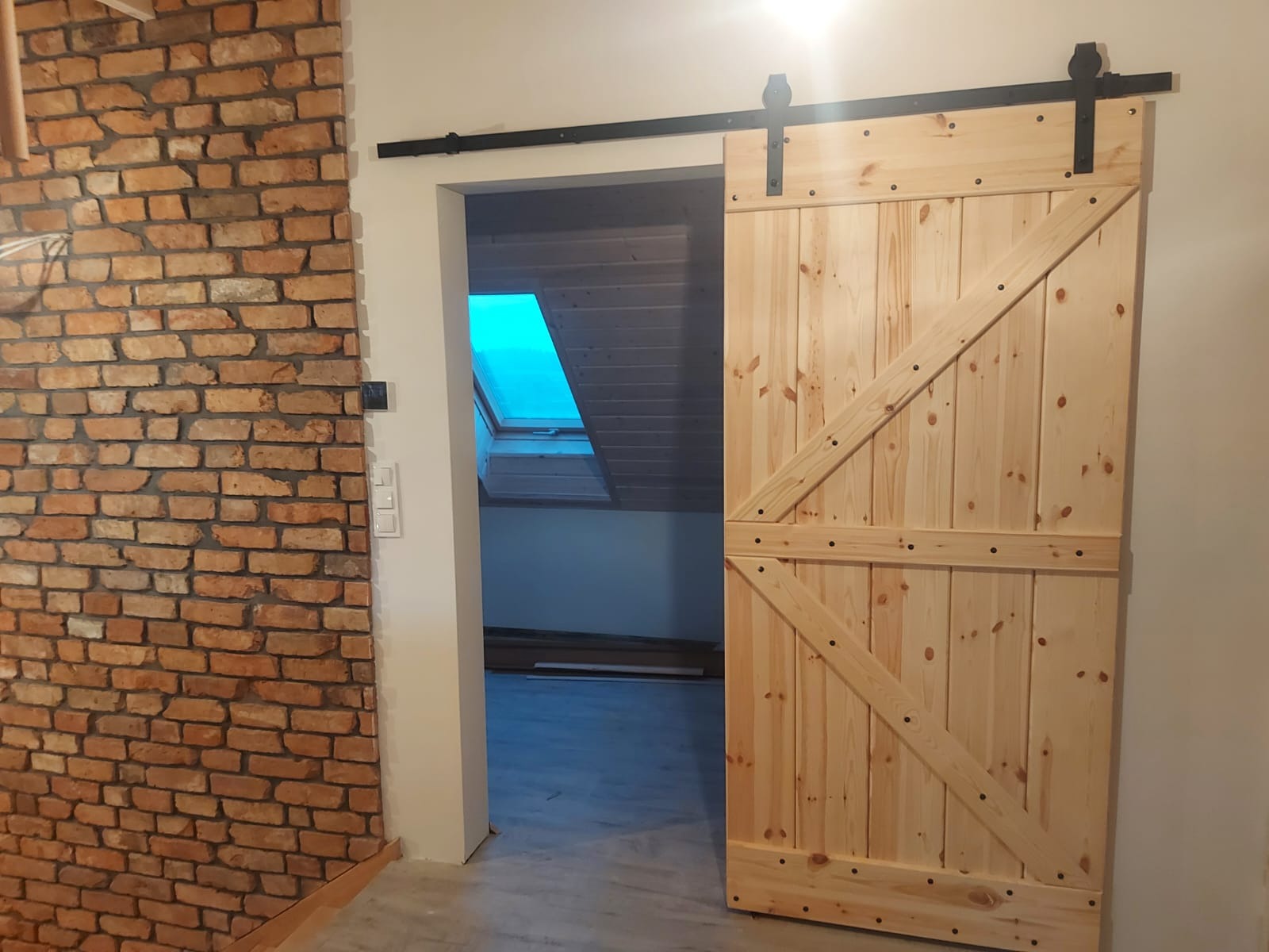 Drewniane drzwi przesuwne w stylu rustykalnym, zamontowane na czarnej szynie, z widoczną ścianą z czerwonej cegły i oknem dachowym w tle.