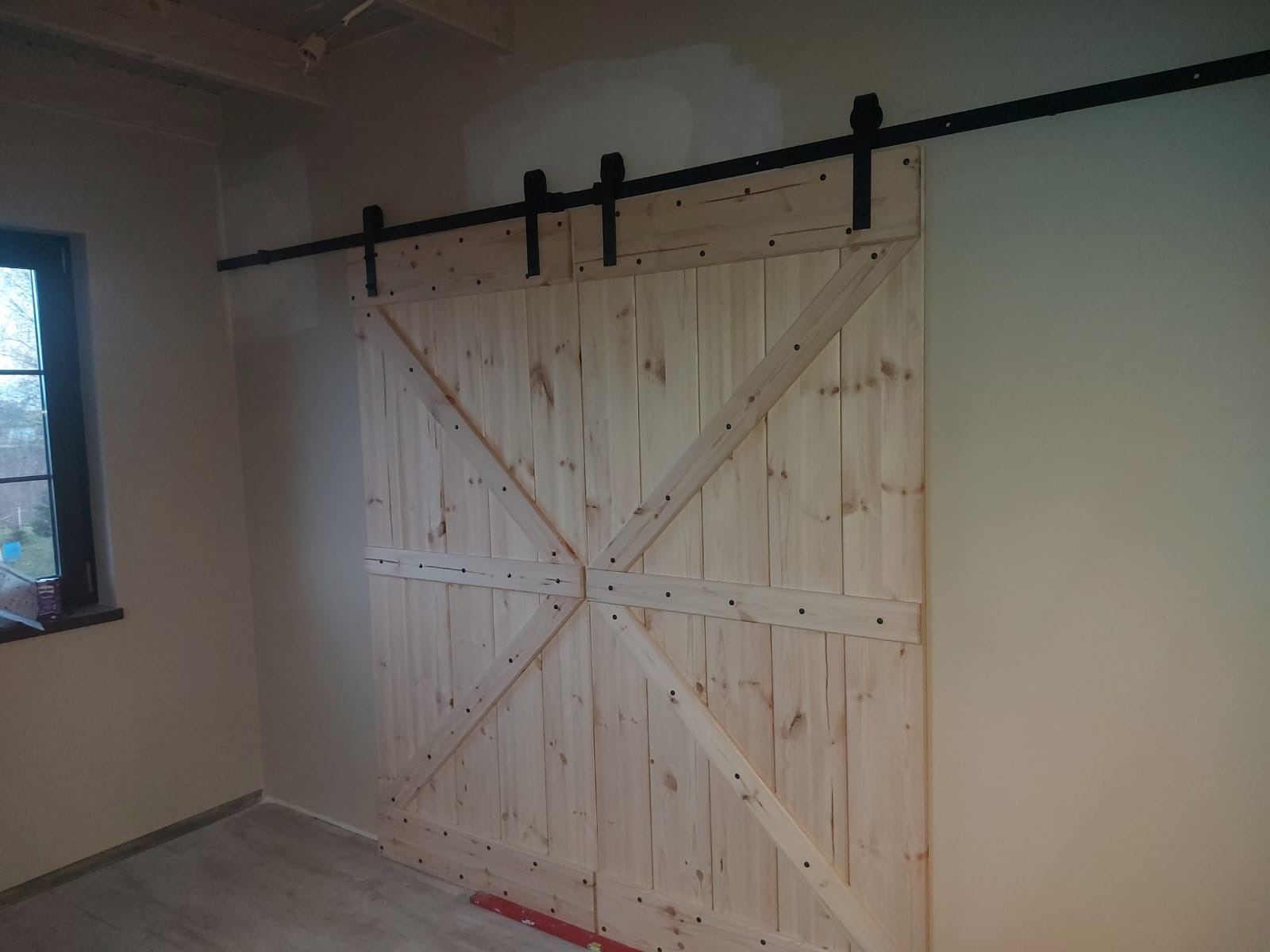 Drewniane drzwi przesuwne w stylu rustykalnym z czarnym systemem szynowym, widoczne poziomica na podłodze przy drzwiach.