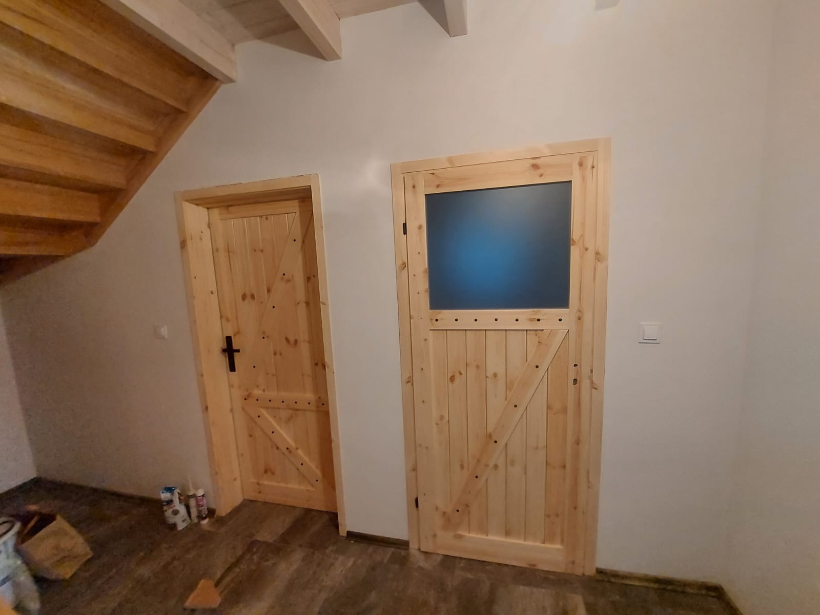 Dwie drewniane drzwi w stylu rustykalnym, jedna z małym, matowym przeszkleniem, druga pełna, z czarną klamką, widoczne drewniane schody i fragment podłogi.