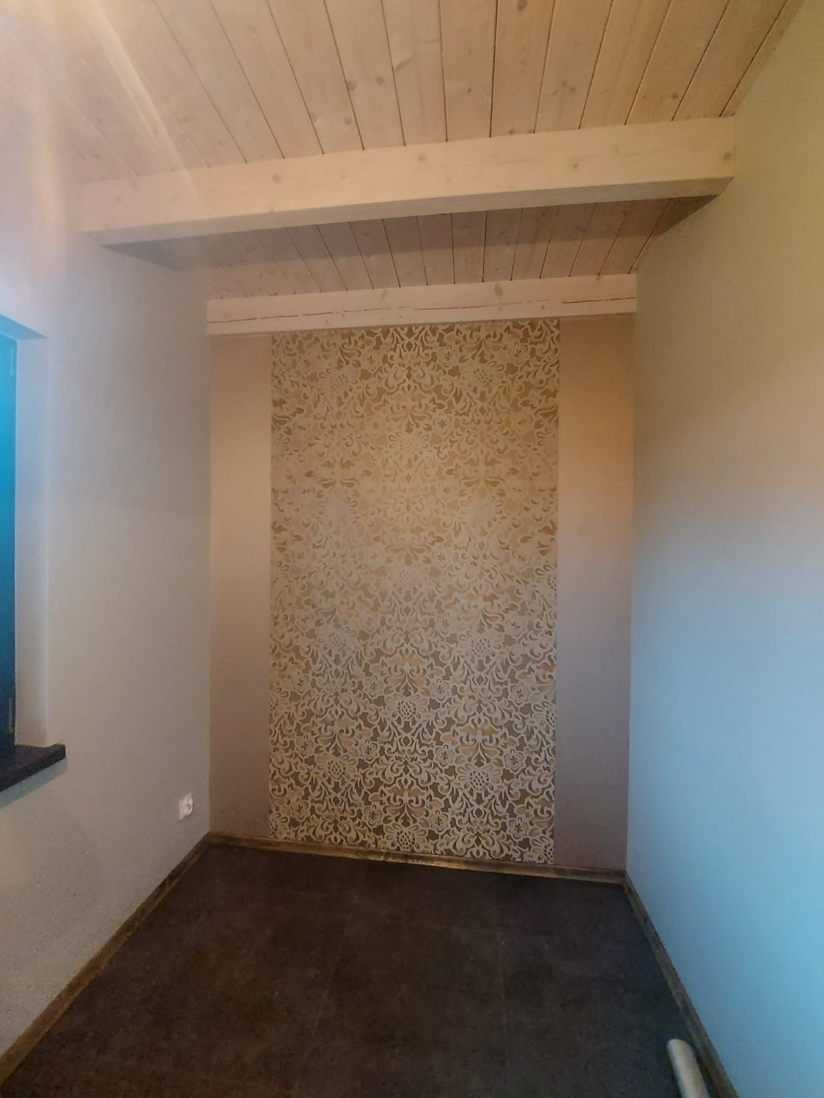 Wykończone wnętrze pokoju z drewnianym sufitem, ścianą z dekoracyjną tapetą w odcieniach beżu i brązową podłogą.