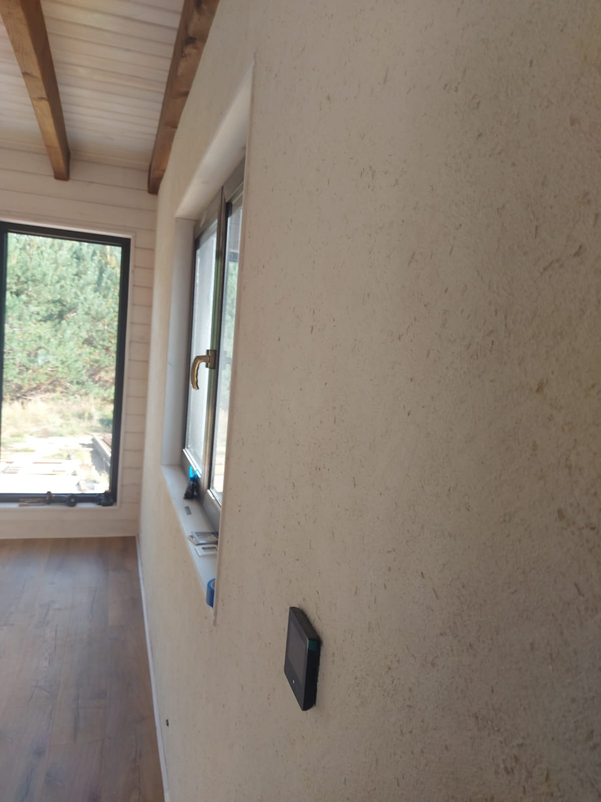 Ściana z fakturą w odcieniach beżu, z zamontowanym panelem sterowania i oknem z widokiem na zieleń, drewniana podłoga i sufit z belkami.