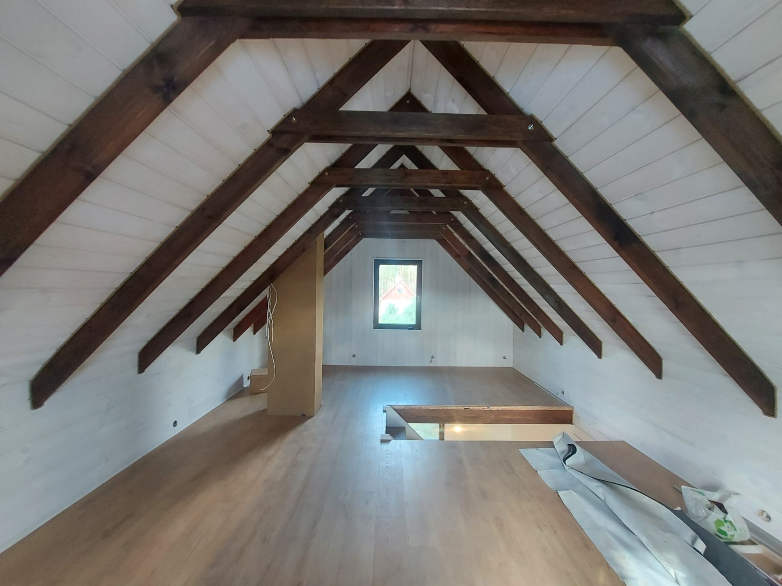 Poddasze po remoncie z drewnianą konstrukcją dachu, wykończone drewnianą boazerią i panelami podłogowymi, widoczne okno dachowe i pionowa zabudowa z przewodami elektrycznymi.
