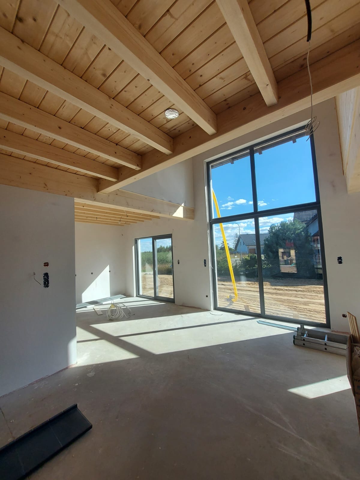 Surowe wnętrze w trakcie prac wykończeniowych: widoczne drewniane belki stropowe, duże okna z widokiem na zewnątrz, surowe ściany, instalacja elektryczna i drabina oparta o okno.