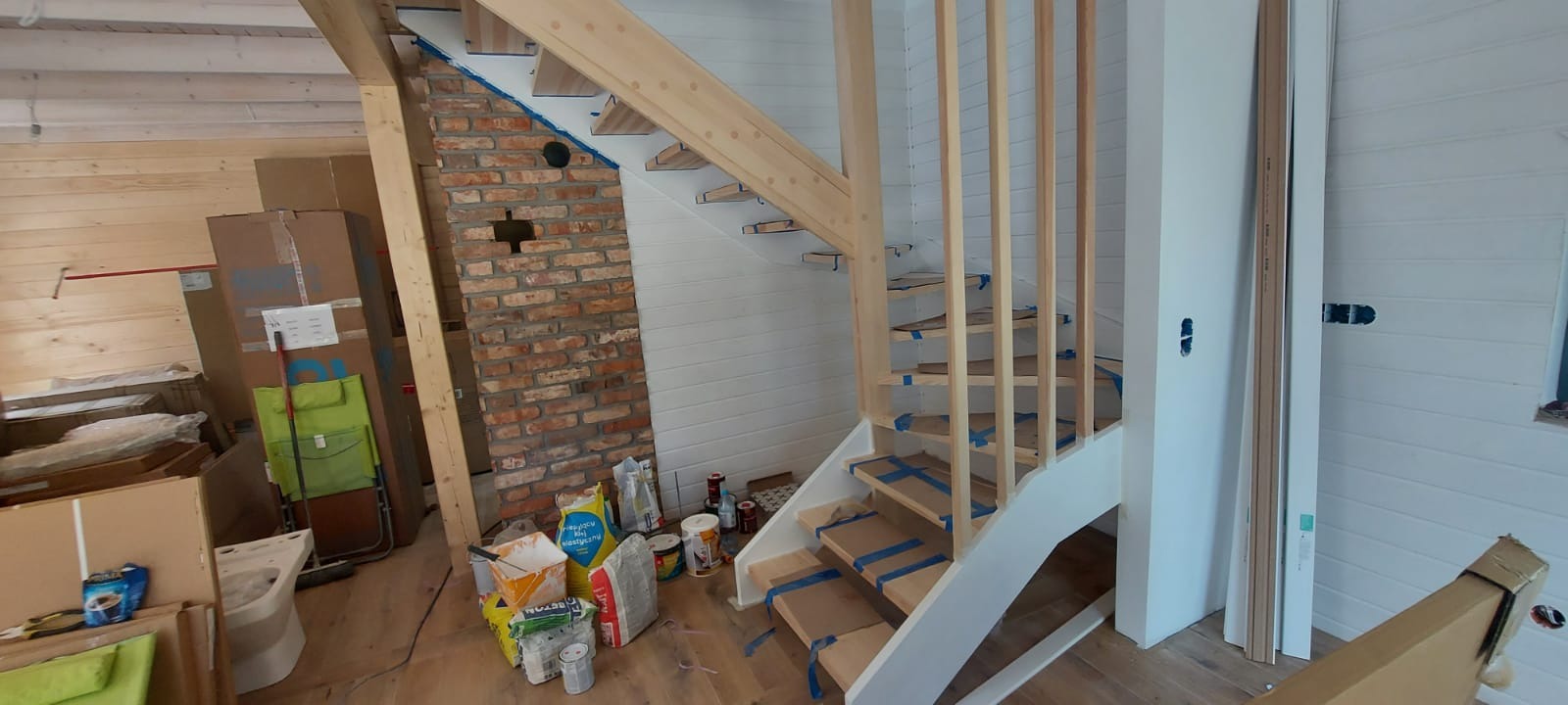Montaż drewnianych schodów ze wstępnie pomalowanymi na biało elementami, zabezpieczonych taśmą malarską, w surowym wnętrzu z boazerią i ceglanym kominem, widoczne materiały budowlane i narzędzia.