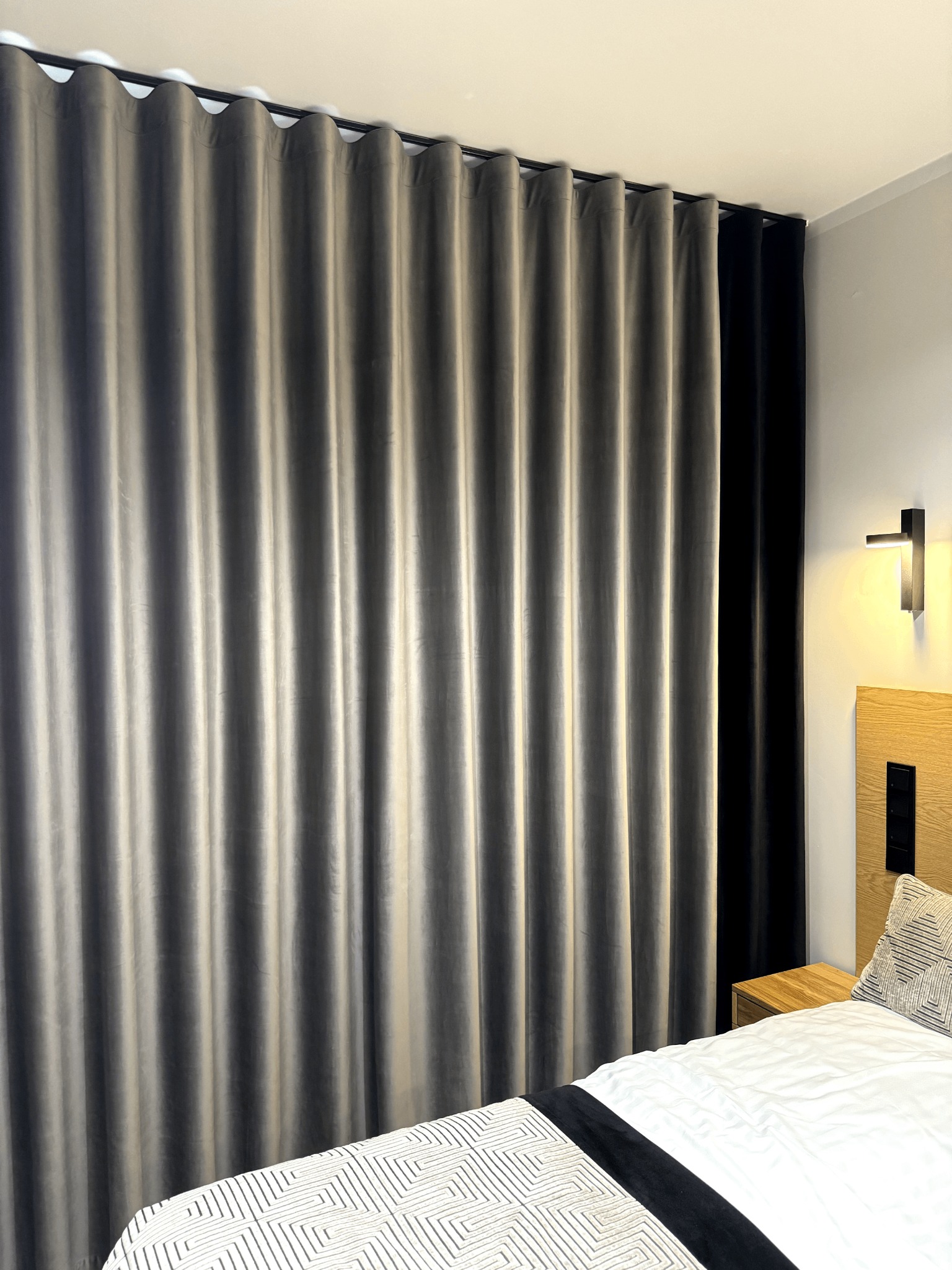 Zasłony zaciemniające w odcieniach szarości i czerni, zawieszone na czarnym karniszu w sypialni, widoczne fragment łóżka z geometrycznym wzorem na narzucie i poduszce, oświetlenie ścienne