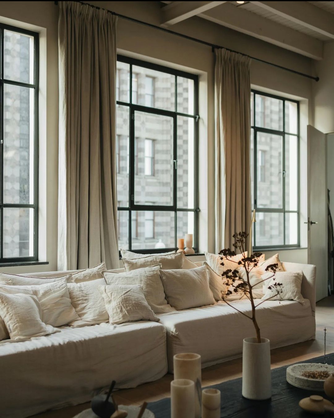 Jasny salon z dużym narożnikiem obłożonym licznymi poduszkami, okna z czarnymi ramami i długimi, beżowymi zasłonami.