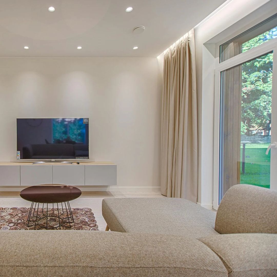 Jasne, nowoczesne wnętrze salonu z beżowymi zasłonami przy oknie, telewizorem na ścianie i częściowo widoczną sofą w neutralnych barwach.