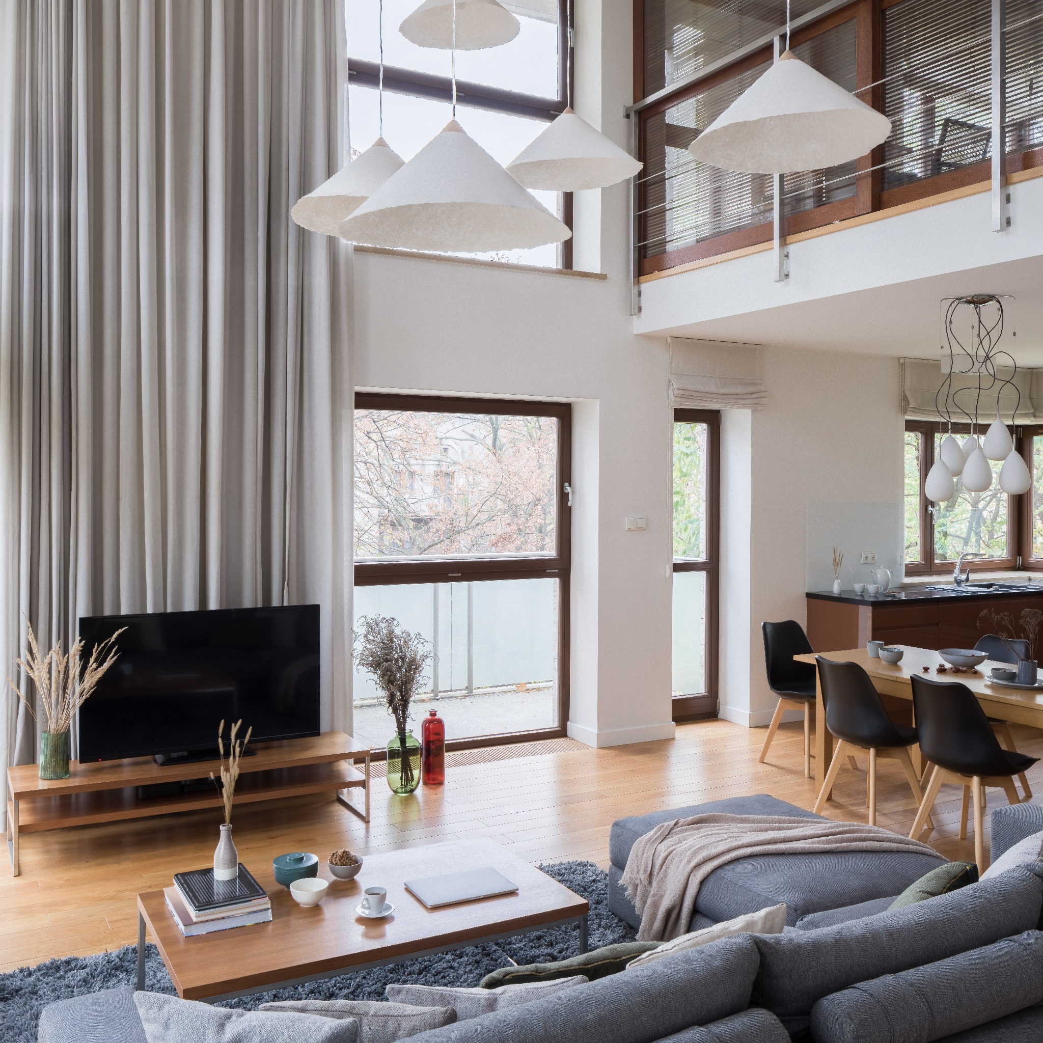 Jasnoszary, długi zasłona w nowoczesnym salonie z wysokim oknem, uzupełniona lampami w kształcie stożków, widoczne także meble: szara sofa, drewniany stolik kawowy i stół jadalny z czarnymi krzesłami.