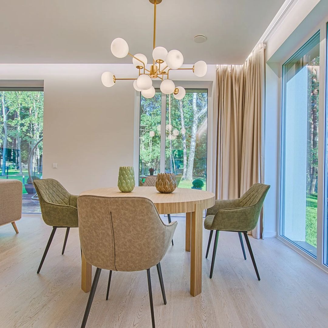Jasne wnętrze jadalni z okrągłym stołem, czterema krzesłami w odcieniach zieleni i beżu, nowoczesną lampą sufitową z białymi kulami oraz widokiem na zieleń za oknem.