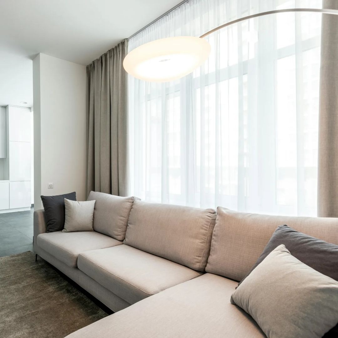 Jasny salon z beżową sofą narożną, szarymi poduszkami i długimi, jasnymi zasłonami w oknie.