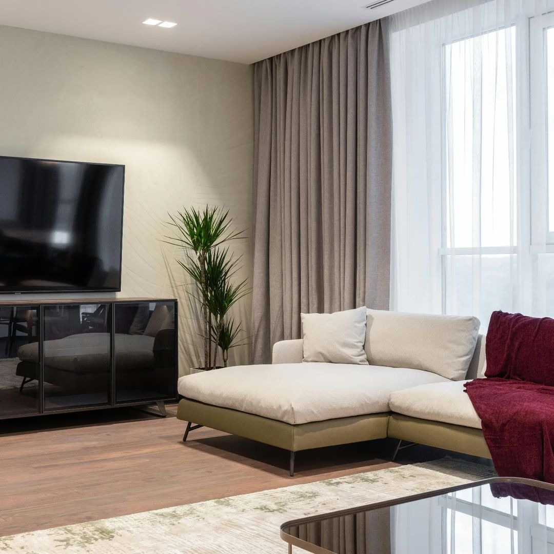 Jasnoszary salon z zasłonami w odcieniach beżu, dużą sofą, rośliną doniczkową i szklanym stolikiem kawowym.