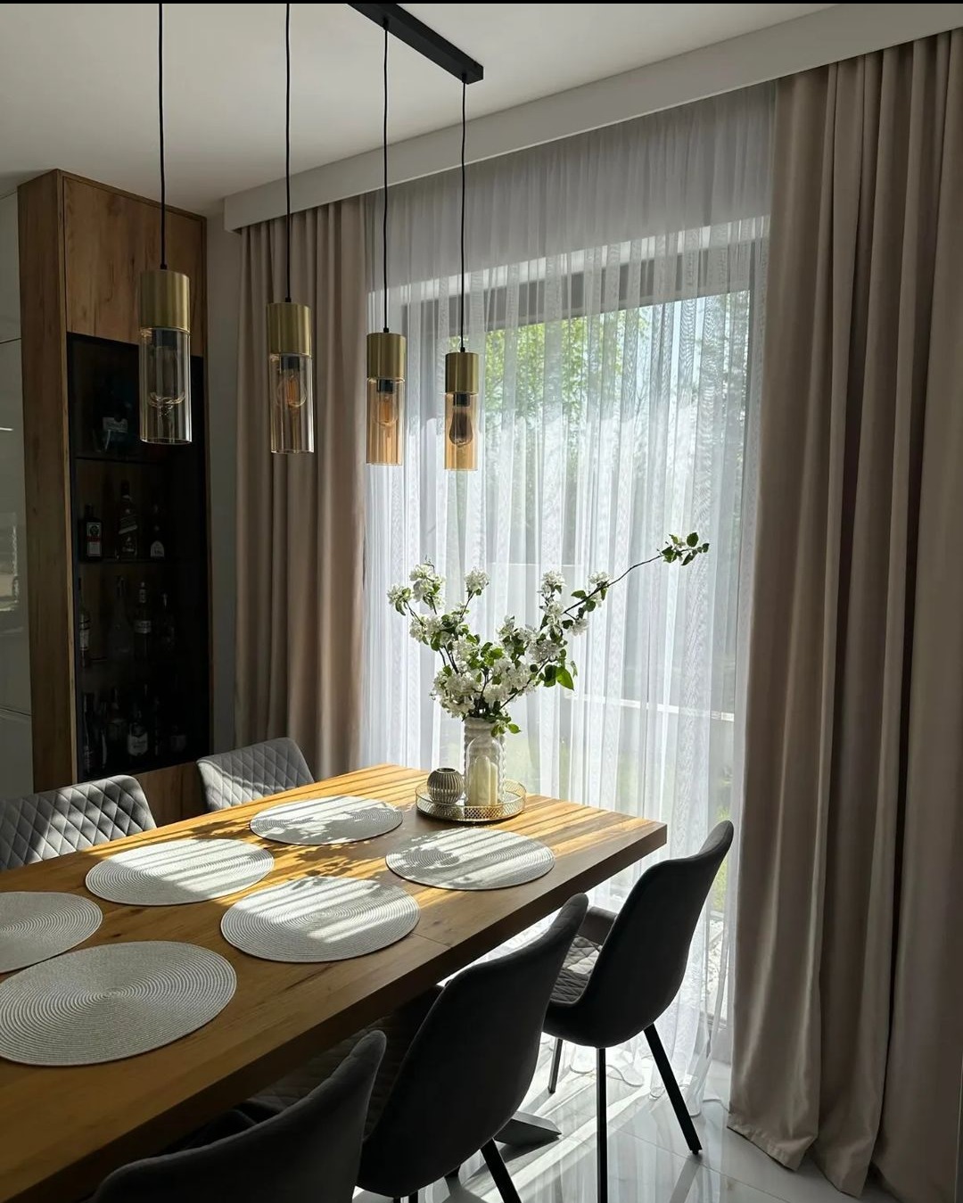 Jadalnia z drewnianym stołem, szarymi krzesłami i beżowymi zasłonami w oknie, oświetlona czterema wiszącymi lampami z elementami złota i szkła.