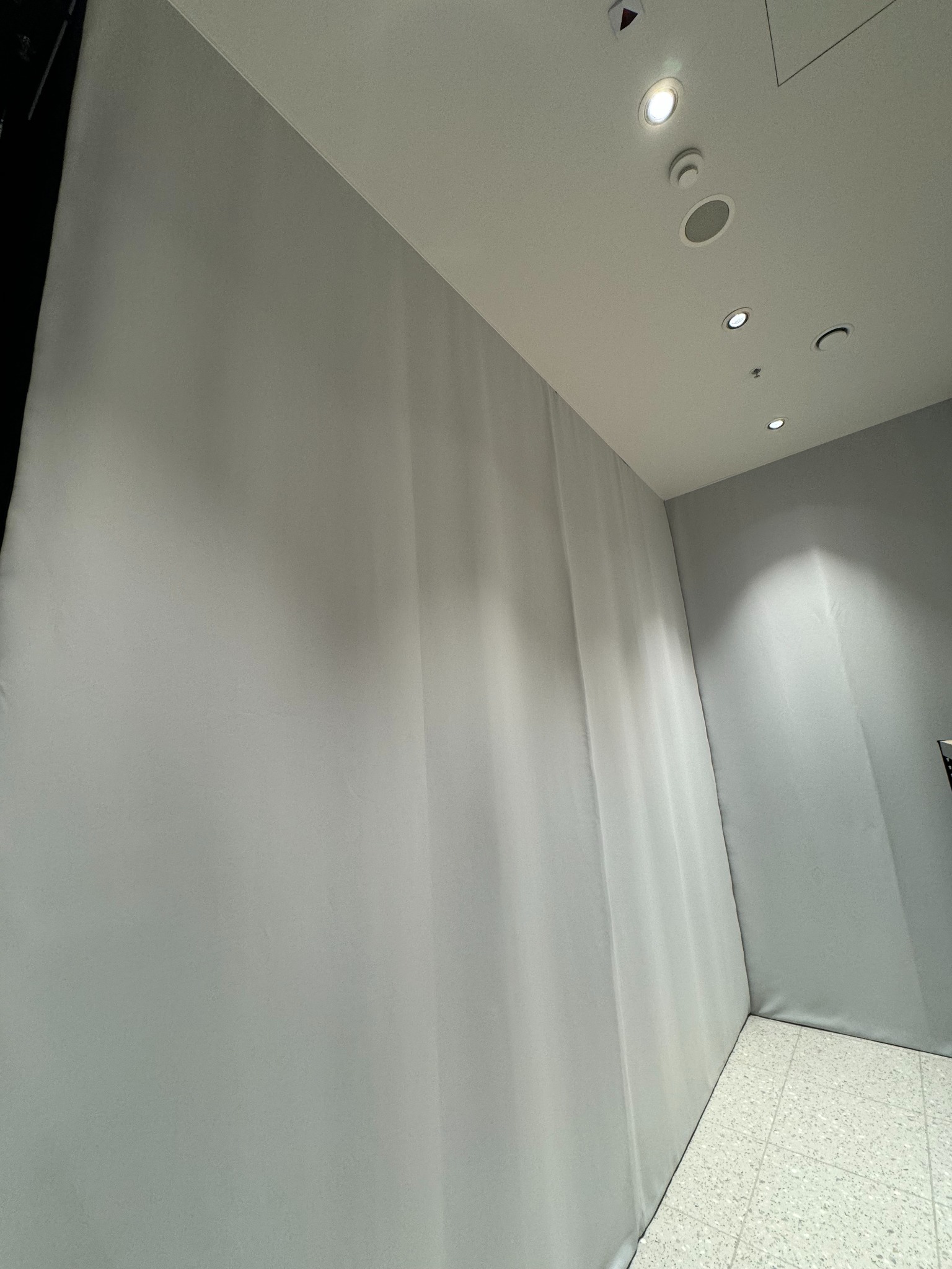 Jasnoszara, matowa tkanina zasłaniająca ścianę w pomieszczeniu z oświetleniem sufitowym punktowym i podłogą z jasnych płytek.