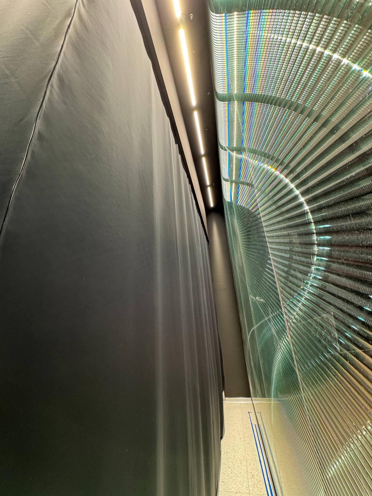 Ciemna, zasłonięta kotara obok futurystycznej, tęczowej instalacji z żaluzji w jasnym pomieszczeniu.