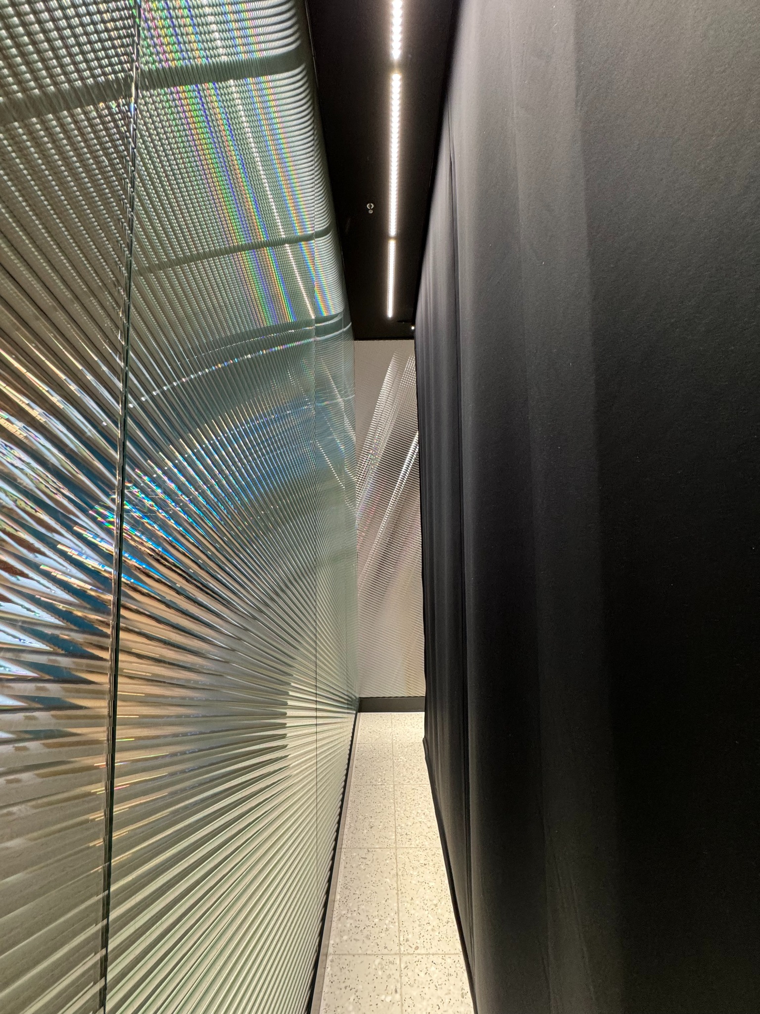 Wąski korytarz z metalową ścianą o prążkowanej fakturze, odbijającej tęczowe światło, oraz czarną, grubą zasłoną po prawej stronie.