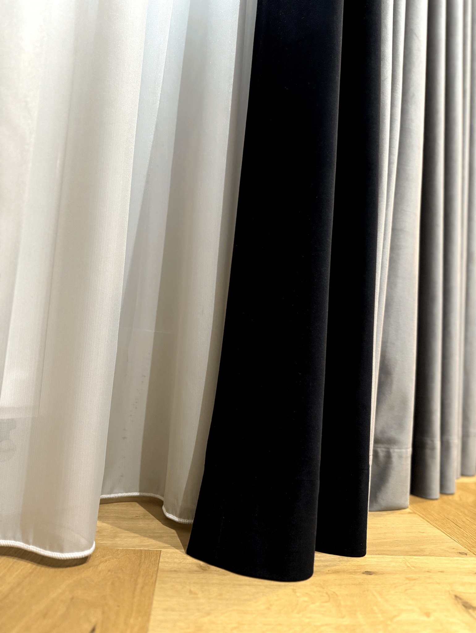 Draperia z różnorodnych tkanin: delikatna, prześwitująca biel, głęboka czerń i stonowana szarość na drewnianej podłodze.
