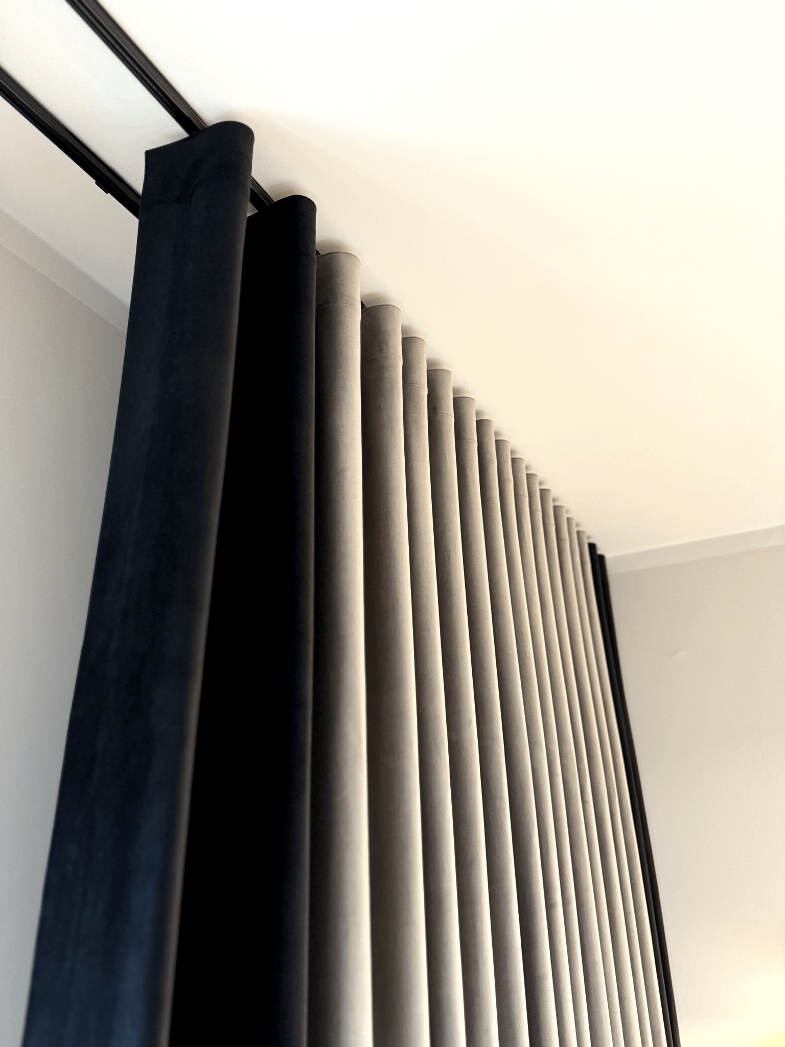 Draperie zaciemniające w odcieniach szarości i czerni, zawieszone na podwójnym karniszu sufitowym, układające się w regularne fałdy.