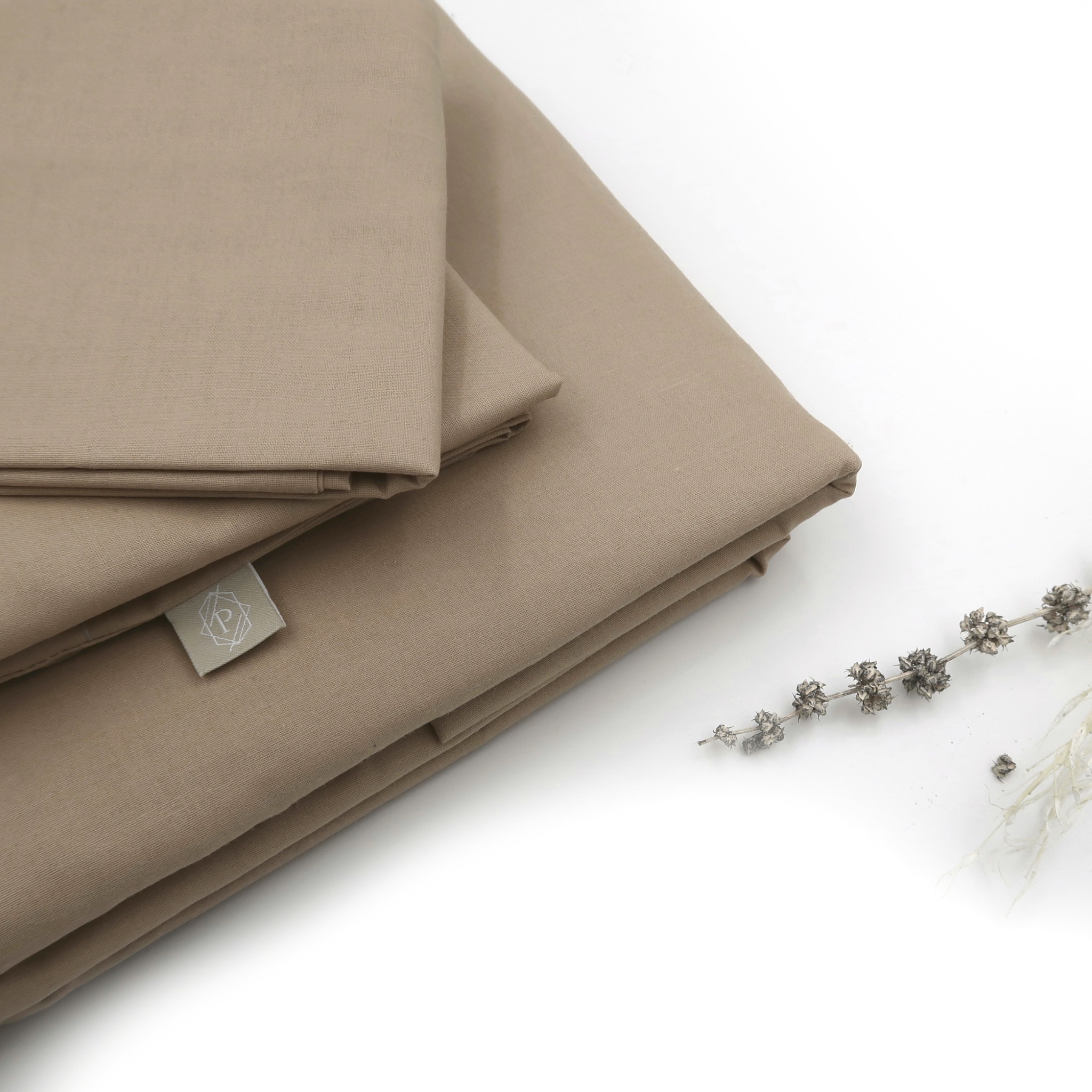 Beżowa pościel bawełniana złożona w kostkę, z widoczną tkaną metką i suszoną gałązką lawendy na białym tle.