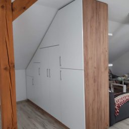 Robert Dzierzgwa R.D.R. - Biała szafa wnękowa na poddaszu w Radoszycach, z drewnianymi elementami wykończeniowymi, dopasowana do skosu dachu, widoczna drewniana belka konstrukcyjna.