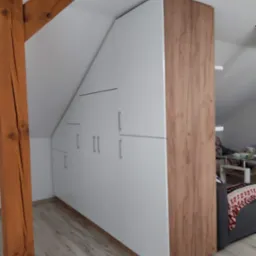 Biała szafa wnękowa na poddaszu w Radoszycach, z drewnianymi elementami wykończeniowymi, dopasowana do skosu dachu, widoczna drewniana belka konstrukcyjna.