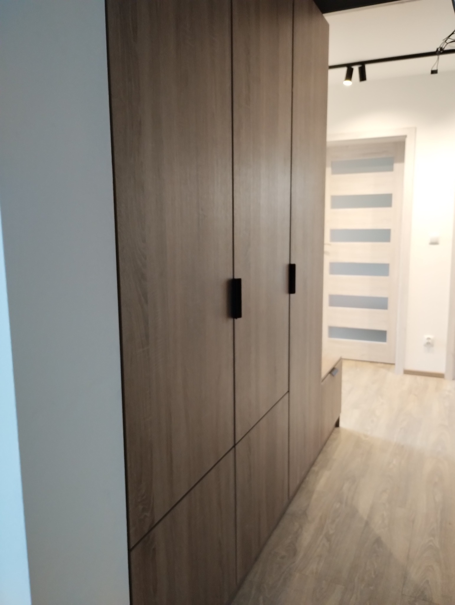 Wysoka szafa w odcieniach drewna z czarnymi uchwytami, umieszczona w korytarzu z panelami podłogowymi i białymi ścianami, widoczna w perspektywie z boku.